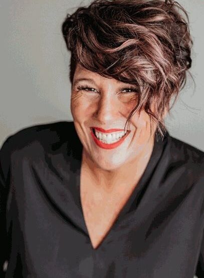 A smiling person with short, wavy, brown hair styled to the side is wearing a black top. They have red lipstick and are posed against a neutral background.