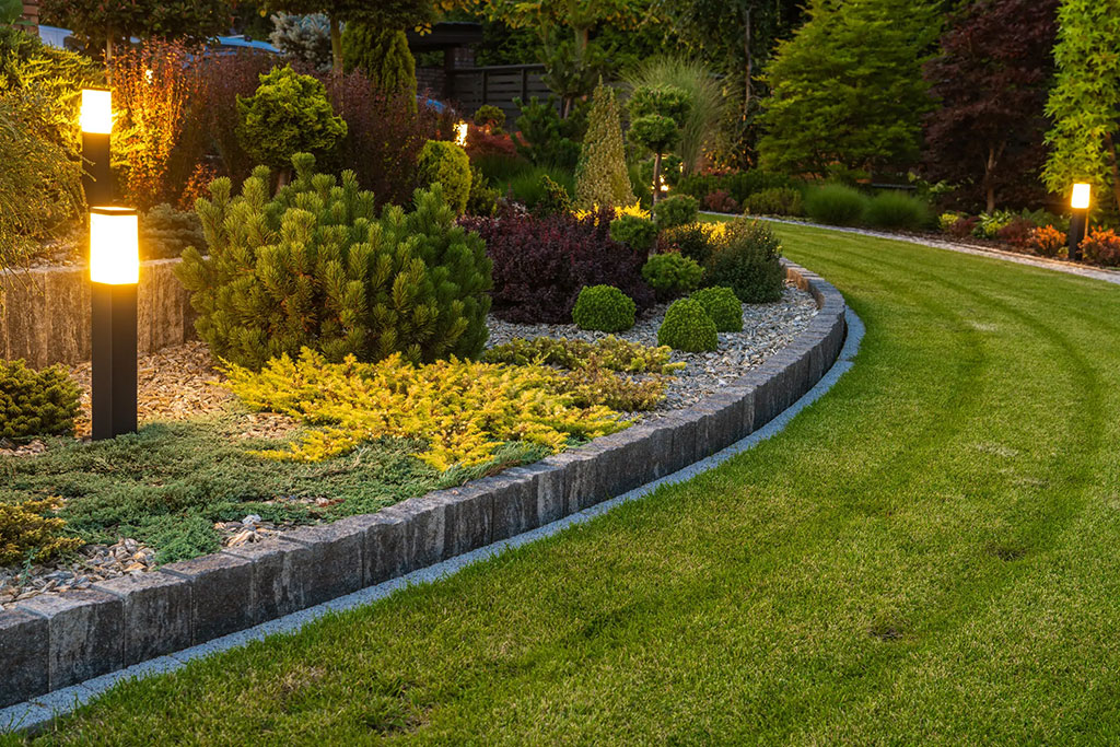 A beautifully landscaped garden with a neatly trimmed lawn and a curved stone-edged flowerbed. The flowerbed is filled with various green shrubs and plants, and illuminated garden lights line the border, casting a warm glow in the evening light.