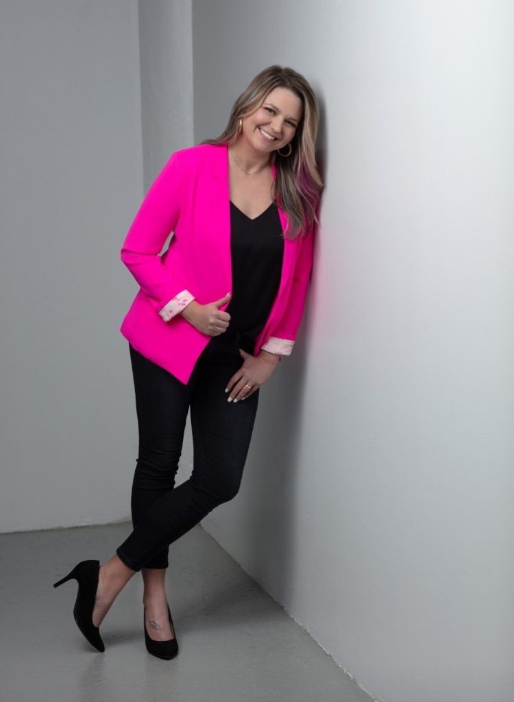 A woman is smiling and leaning against a white wall. She is wearing a bright pink blazer over a black top, dark jeans, and black high heels. Her long hair is styled straight, and she has one hand in her blazer pocket. The background is simple and muted.