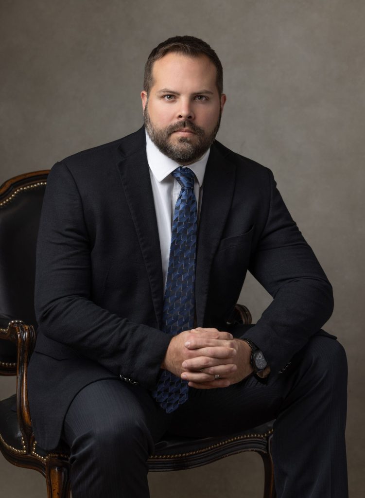 A man with short hair and a beard wearing a dark suit, white dress shirt, and blue patterned tie sits on a chair with his hands clasped. The background is plain and neutral-toned.